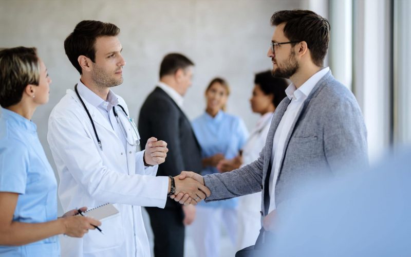 I’m glad we made an agreement! Male doctor handshaking with a businessman while meeting in a hallway at medical clinic.